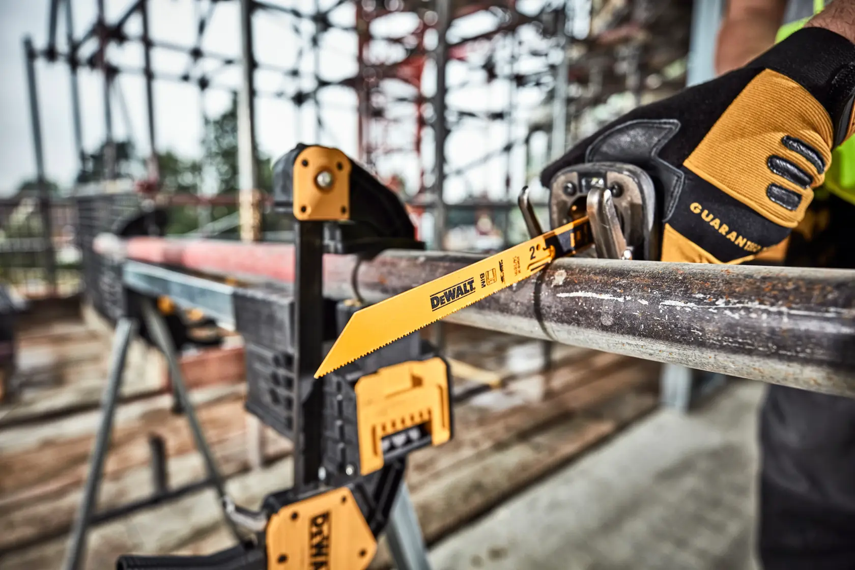 Une lame de scie sabre DEWALT DT2416 coupe un tuyau en métal sur un chantier de construction. Le tuyau est fixé avec des serre-joints et une personne portant des gants de travail utilise la scie.