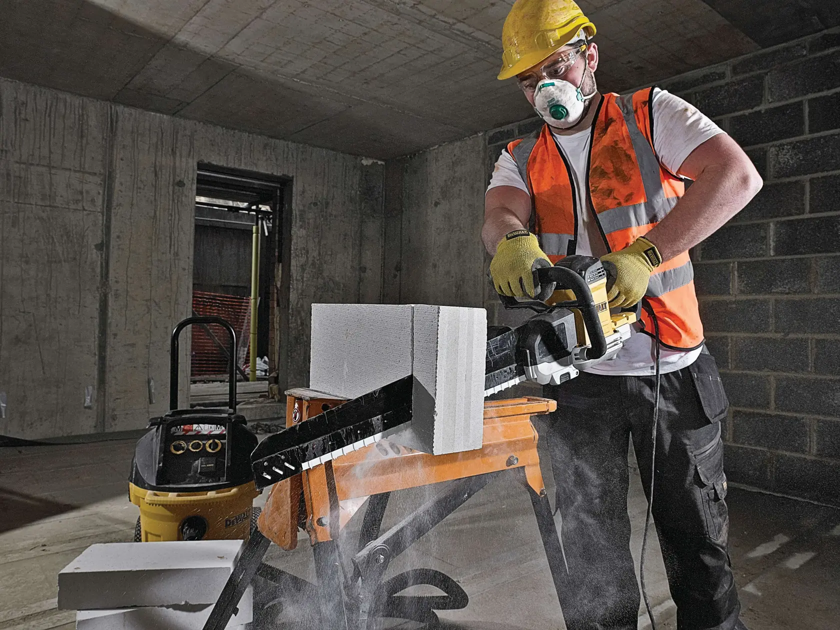 Ouvrier coupe un bloc de béton avec un outil DEWALT en intérieur, gilet, gants et casque de sécurité.