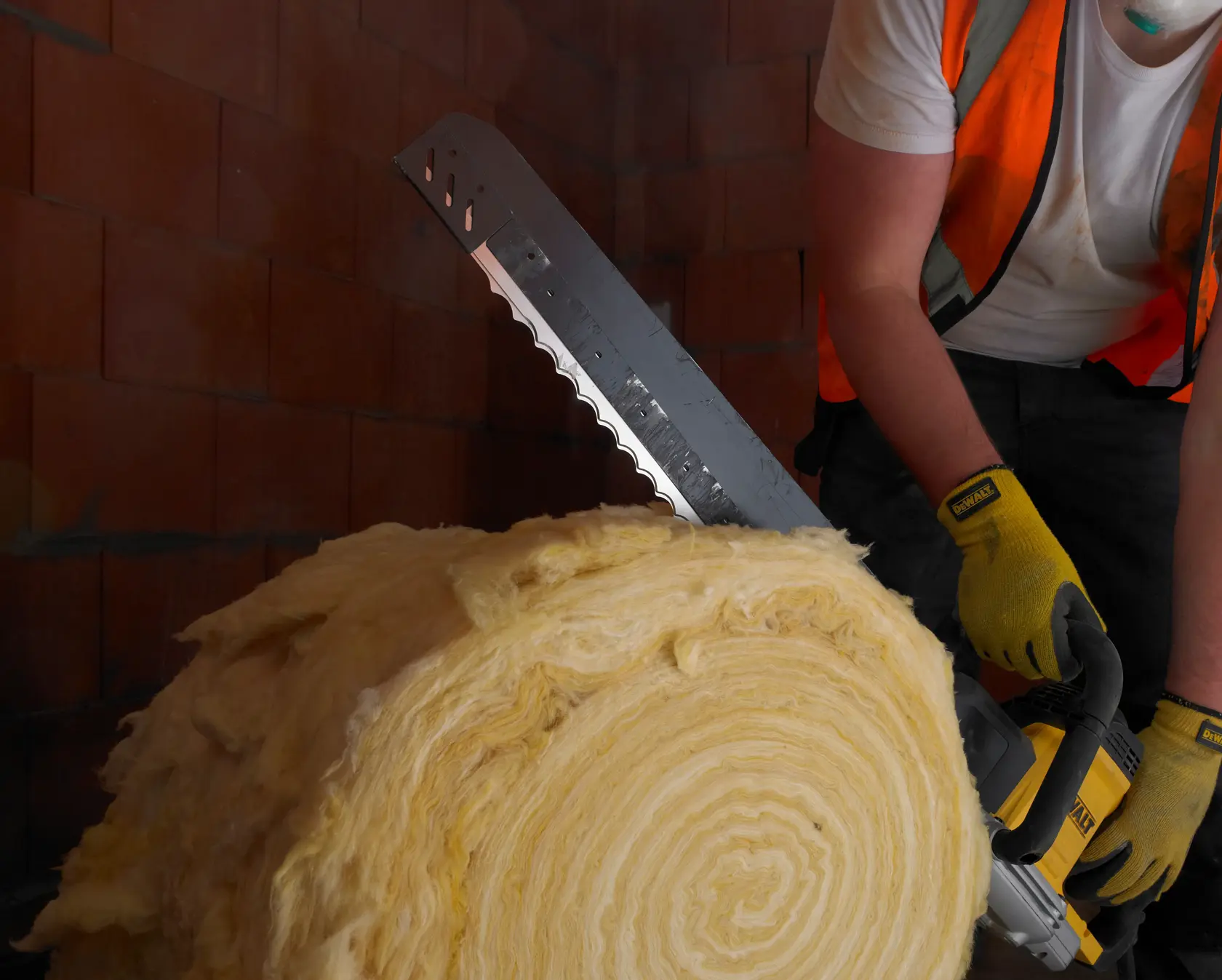 Personne découpe un rouleau d’isolant avec une scie DEWALT, portant gants et gilet dans une zone en briques.