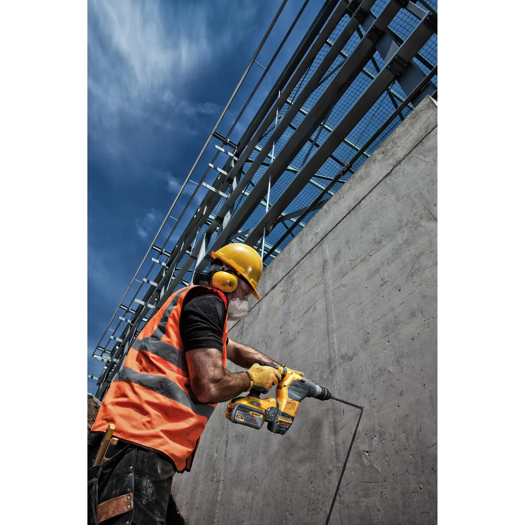 Un ouvrier du bâtiment portant un casque jaune, une protection auditive, un gilet de sécurité et des gants utilise une perceuse DEWALT sur un mur en béton sur un chantier sous un ciel bleu.