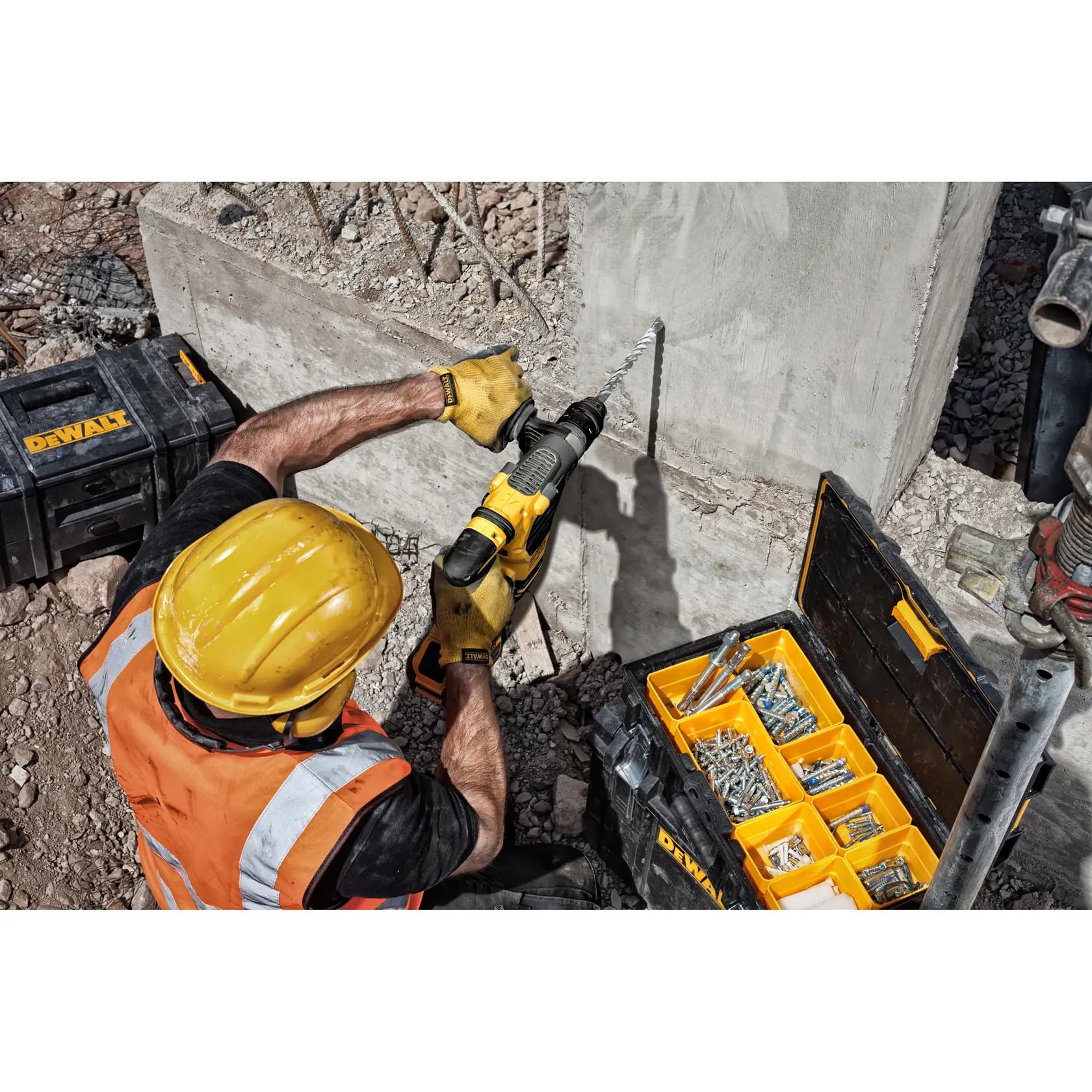 Un ouvrier du bâtiment portant un casque jaune et un gilet de sécurité orange utilise une perceuse DEWALT avec un long foret pour travailler sur un mur en béton. À côté de lui se trouvent des boîtes à outils DEWALT, dont une avec des compartiments organisés contenant diverses vis et fixations.