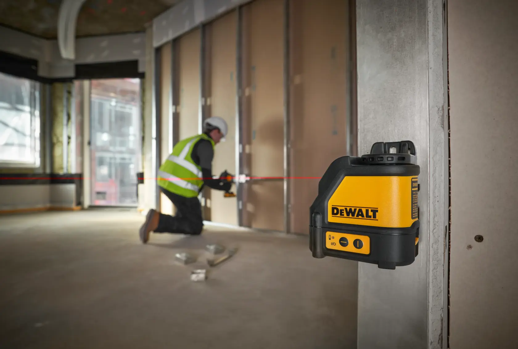 Un niveau laser DEWALT DW088K est fixé sur une colonne métallique, projetant une ligne horizontale rouge à travers l'intérieur d'un chantier. En arrière-plan, un ouvrier portant un gilet de sécurité et un casque aligne des matériaux le long de la ligne laser.