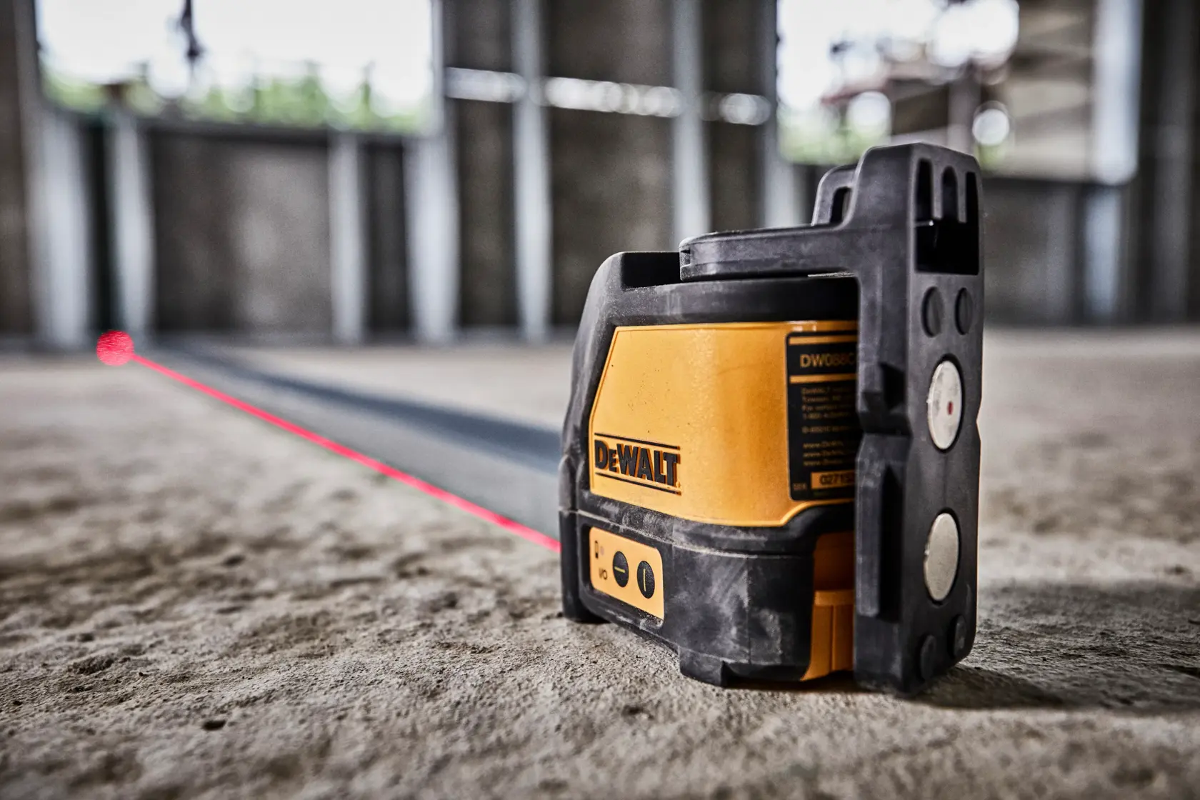 Un outil laser à lignes croisées auto-nivelant DEWALT DW088K projette une ligne laser rouge sur le sol à l'intérieur d'un bâtiment inachevé avec des murs en béton.