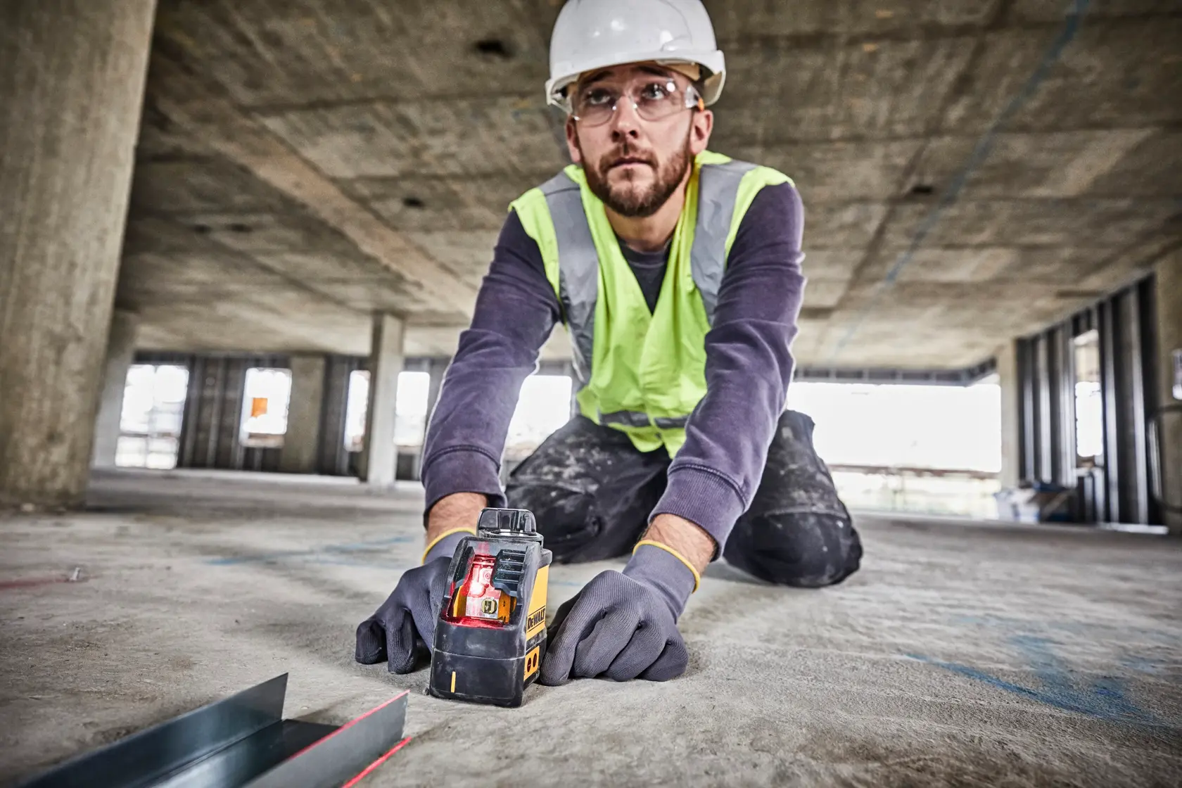 Une personne portant un casque, un gilet de sécurité et des gants est agenouillée sur le sol d'un chantier de construction, utilisant un niveau laser DEWALT DW088K. L'appareil projette une ligne laser rouge visible pour l’alignement. Les environs montrent un bâtiment non terminé avec des colonnes de béton et des fenêtres ouvertes.