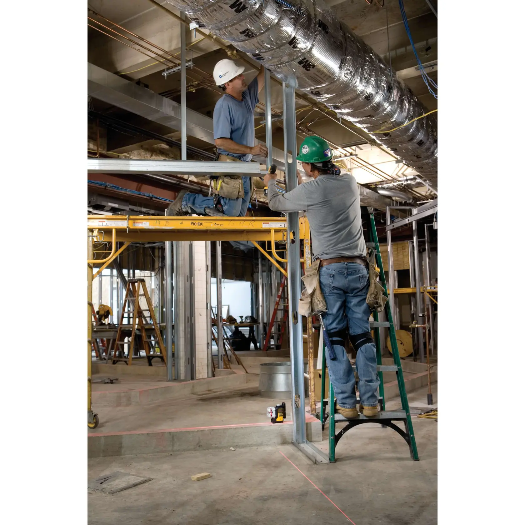 A DEWALT DW089K laser level is positioned on the ground at a construction site while a worker wearing a hard hat and tool belt measures with a tape measure on the floor. The background shows various construction equipment, ladders, and exposed ceiling pipes.