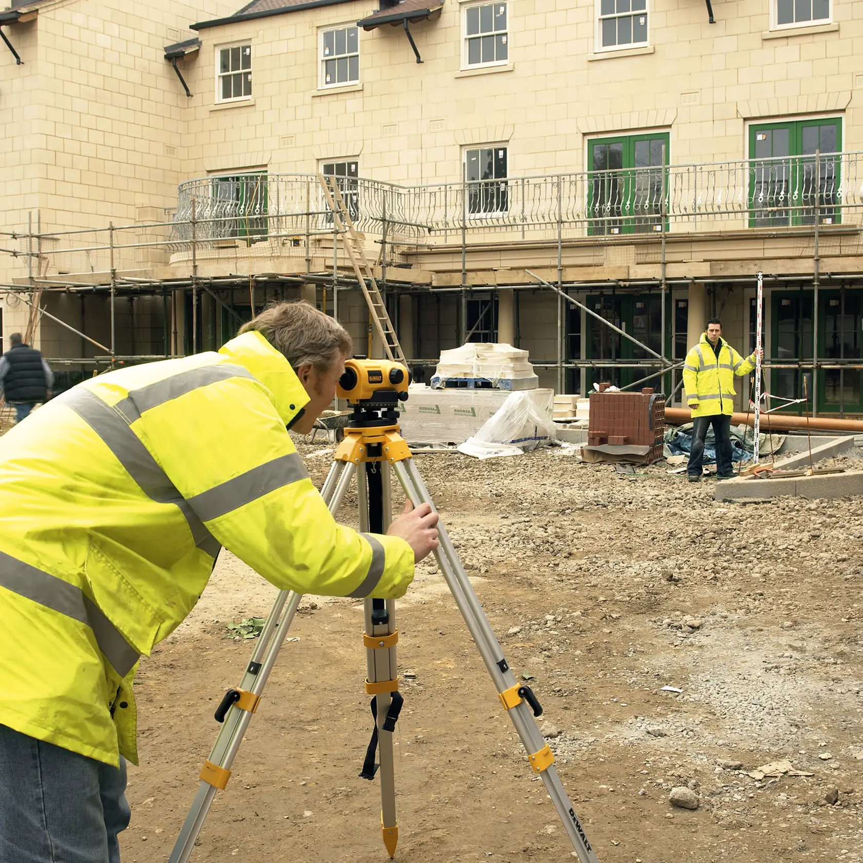 Ouvrier utilise le trépied DEWALT DW096PK sur chantier, un autre tient une tige de mesure.