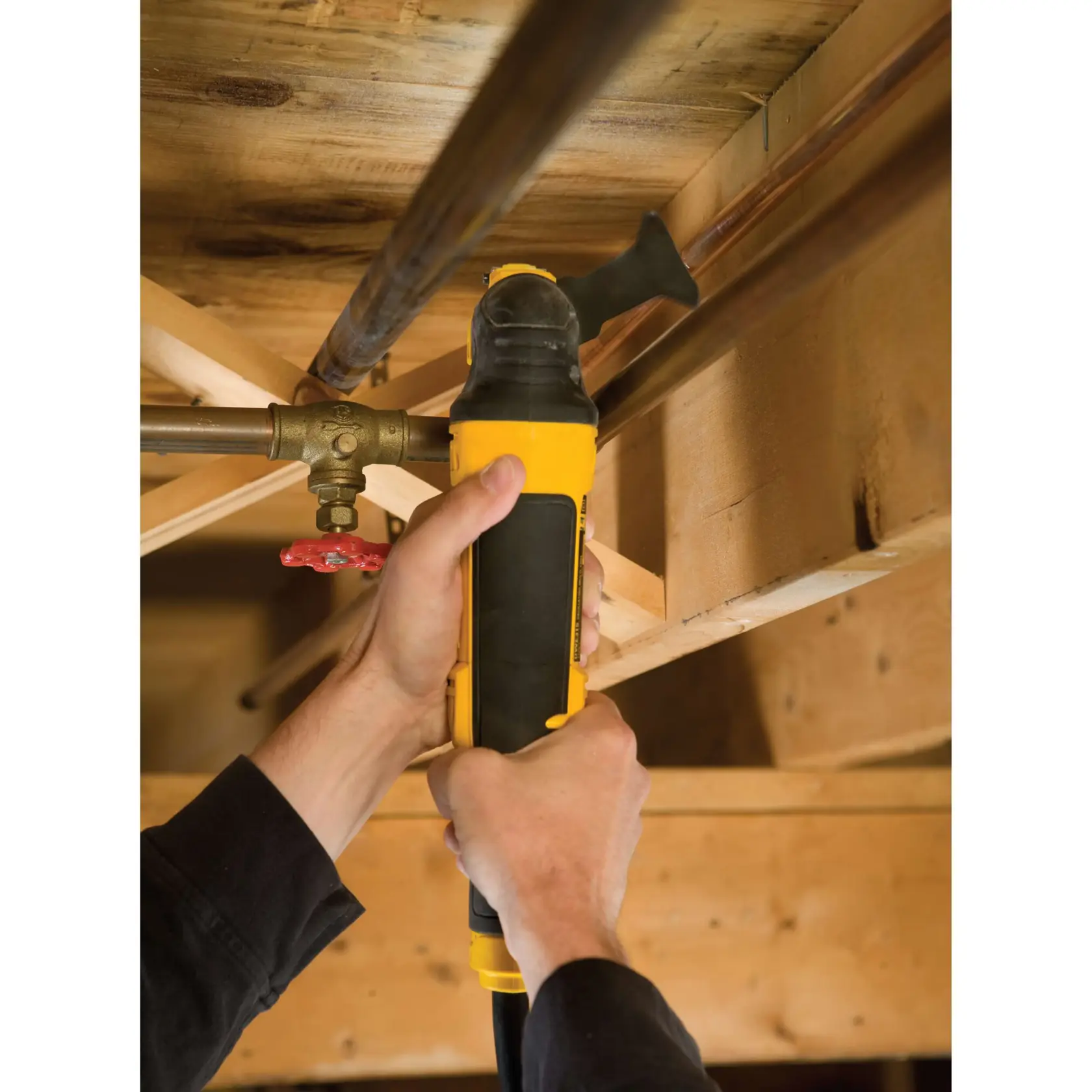 Mains utilisant un outil DEWALT sur un tuyau sous des poutres en bois. Outil jaune et noir.