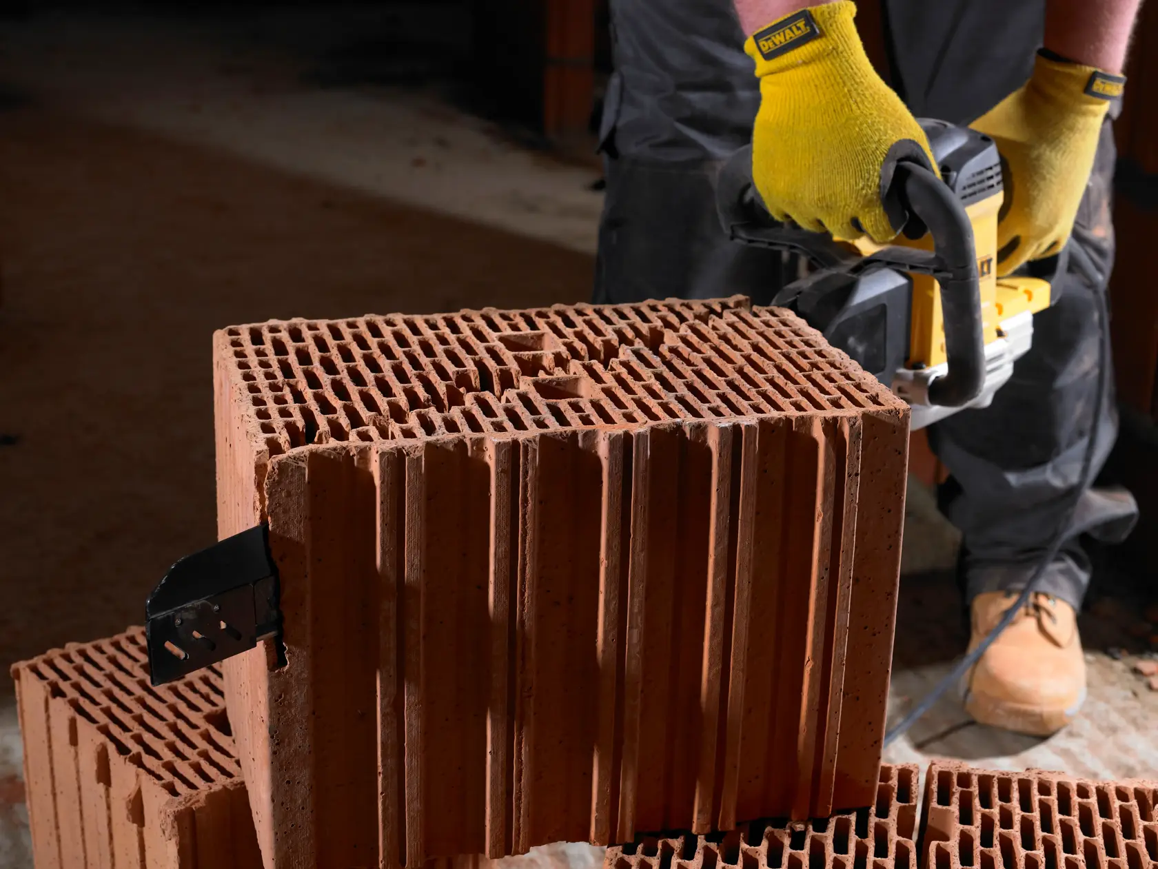 Une personne portant des gants jaunes utilise un outil électrique DEWALT pour découper une grande brique creuse en argile. L'outil est placé contre la brique et la personne se tient sur une surface poussiéreuse.