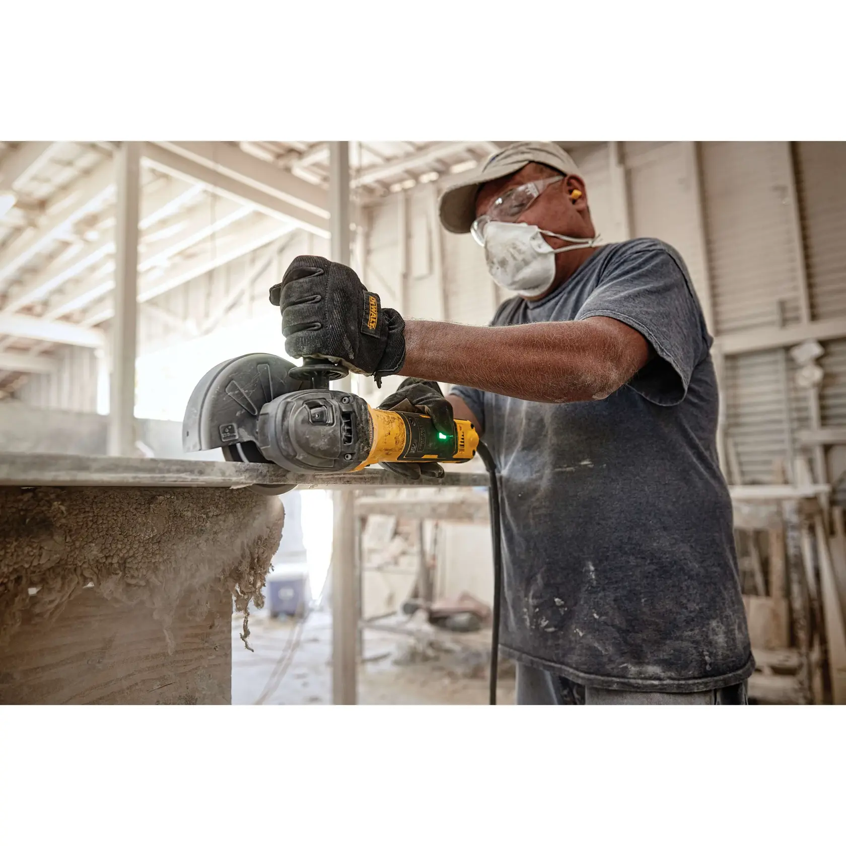Un ouvrier portant des équipements de protection, dont des gants, un masque, des lunettes de sécurité et des bouchons d'oreilles, utilise une meuleuse d'angle DEWALT pour couper un matériau dans un atelier industriel.