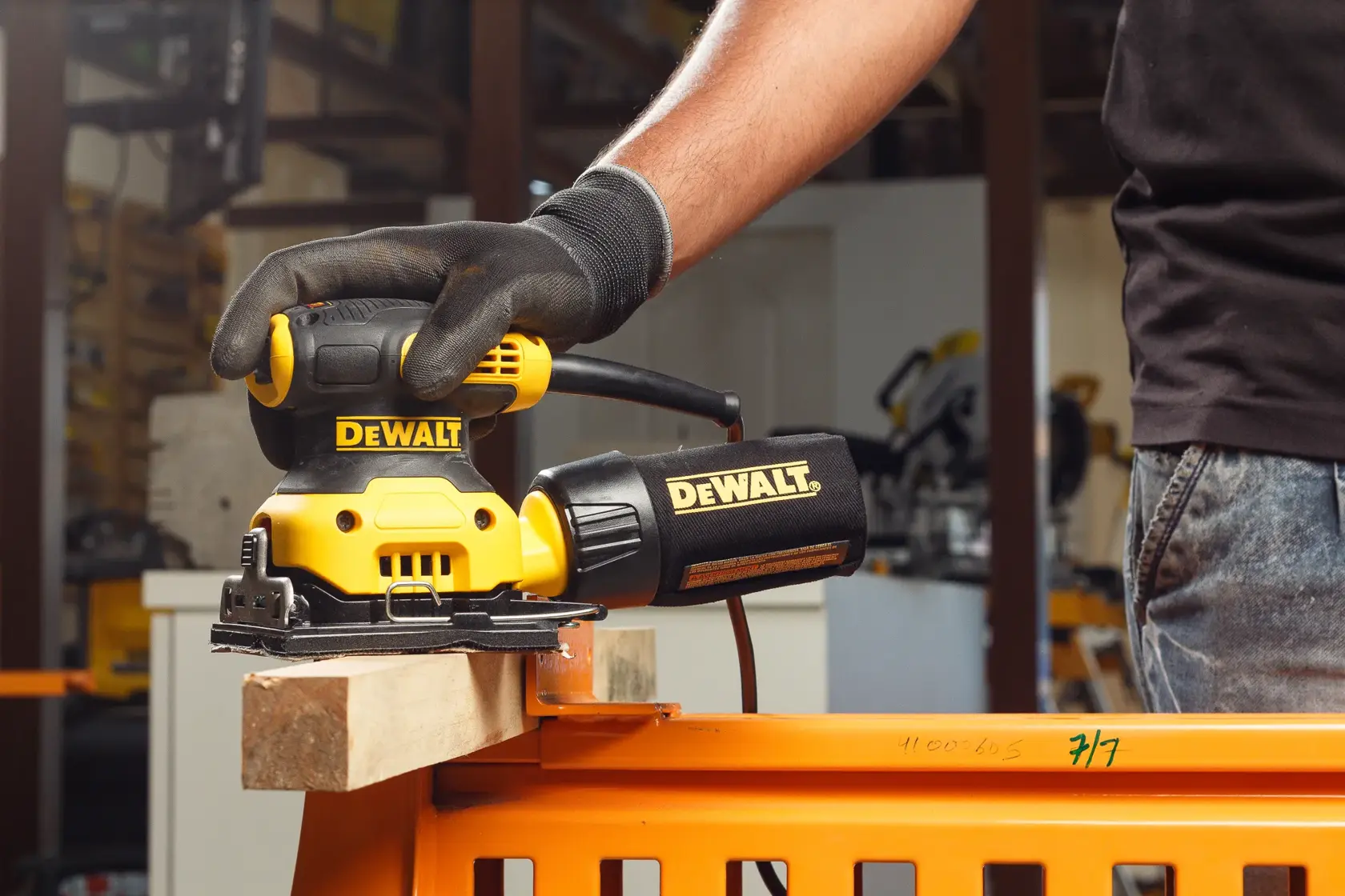 Vue rapprochée d'une main gantée utilisant une ponceuse électrique jaune et noire DEWALT DWE6411 sur une surface en bois à l'extérieur.