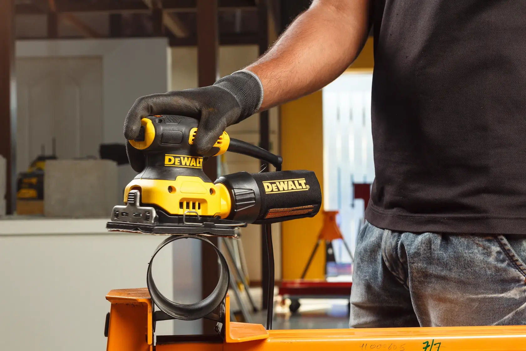 Gros plan d'une personne portant des gants utilisant une ponceuse électrique jaune DEWALT pour lisser la surface d'une planche en bois.