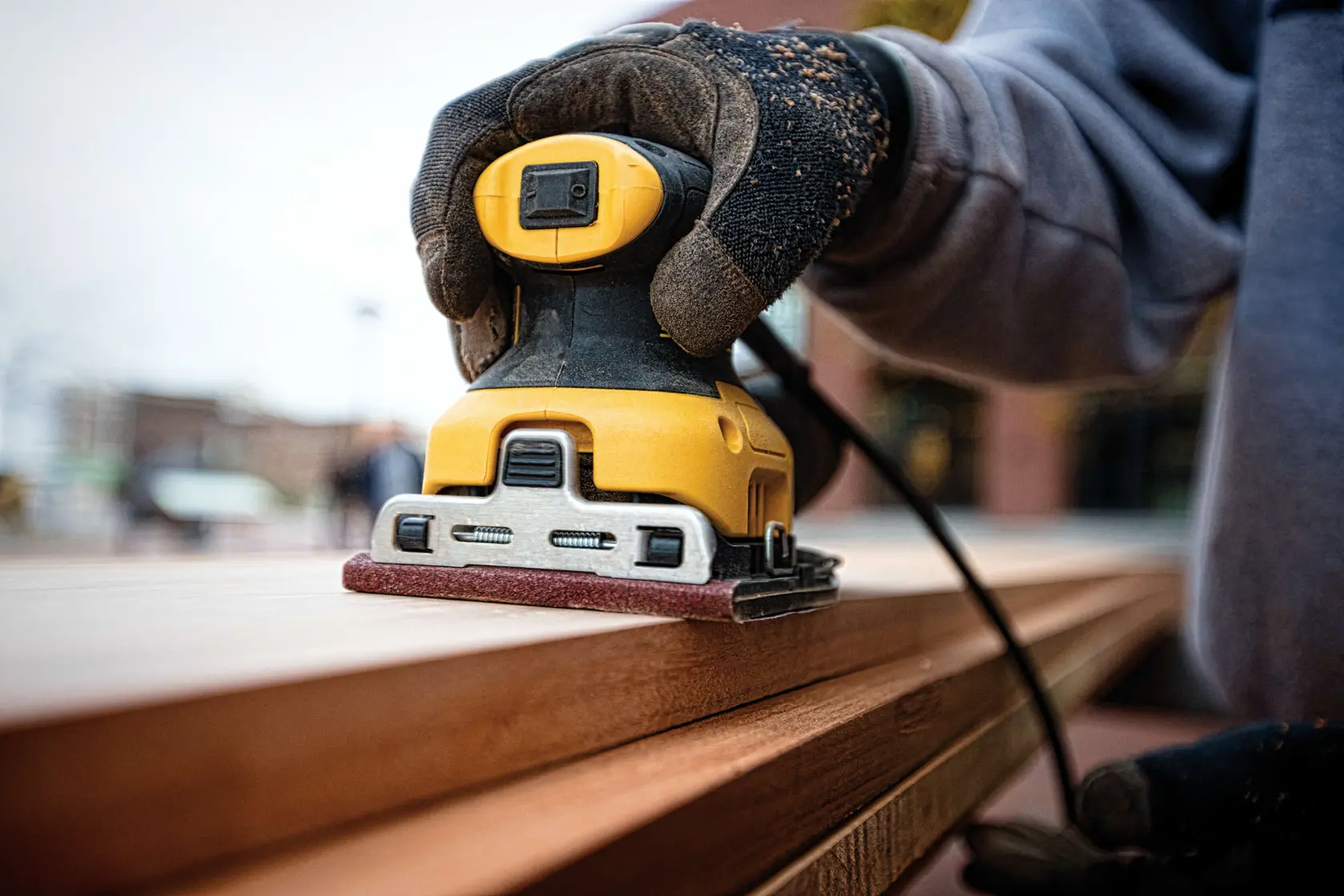 Une personne utilise une ponceuse électrique DEWALT DWE6411 sur une surface en bois à l'intérieur d'un atelier, la ponceuse étant tenue verticalement avec le sac à poussière attaché.