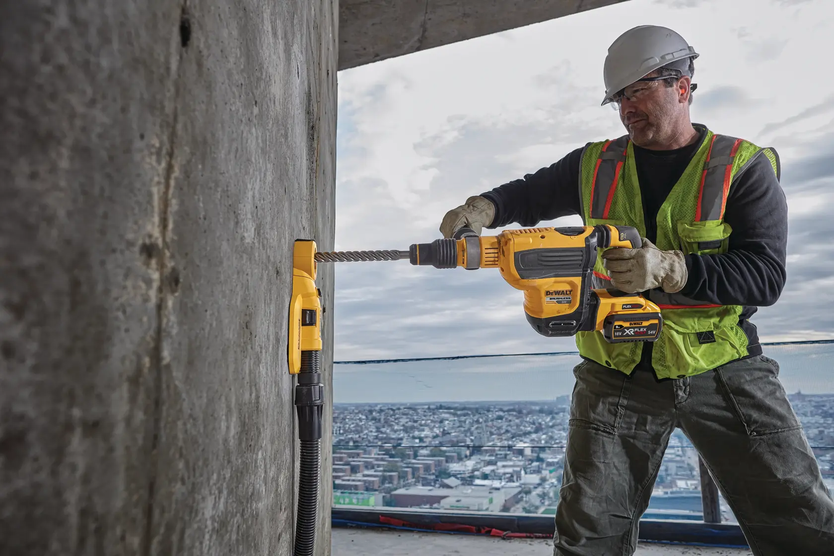 Un ouvrier du bâtiment utilisant une perceuse DEWALT avec un accessoire d'extraction de poussière (SKU : DWH050) pour percer un mur en béton, équipé d'un casque, de gants et d'un gilet haute visibilité, avec une vue sur la ville en arrière-plan.