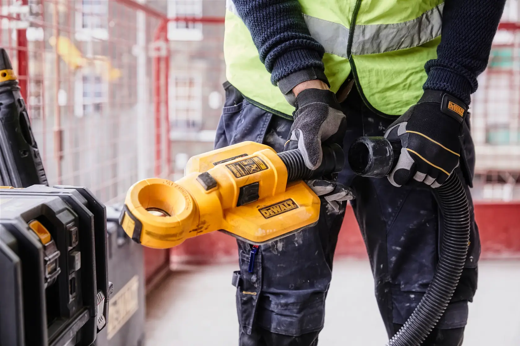 Une personne portant un gilet de sécurité et des gants utilise un accessoire d'aspiration de poussière DEWALT DWH050 sur un chantier. L'outil jaune est connecté à un tuyau et placé près d'un coffre de rangement DEWALT.