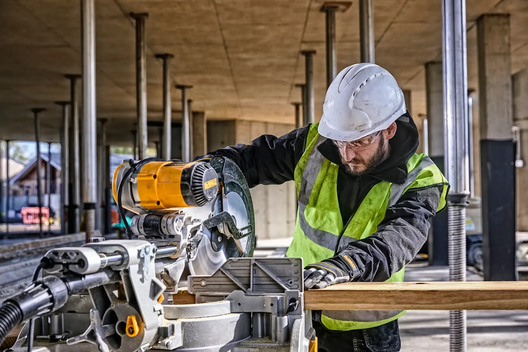 Un ouvrier de construction portant un casque et un gilet haute visibilité utilise une scie à onglets DEWALT DWS780 sur un chantier. La scie coupe une pièce de bois, et des colonnes métalliques ainsi que des matériaux de construction sont visibles en arrière-plan.