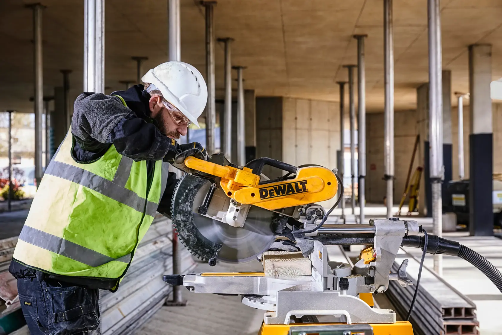 Une scie à onglet DEWALT DWS780 utilisée pour couper une planche en bois sur un chantier de construction. L’opérateur porte un casque de sécurité, des gants et un gilet haute visibilité. L’arrière-plan montre un bâtiment en cours de construction avec des piliers verticaux.