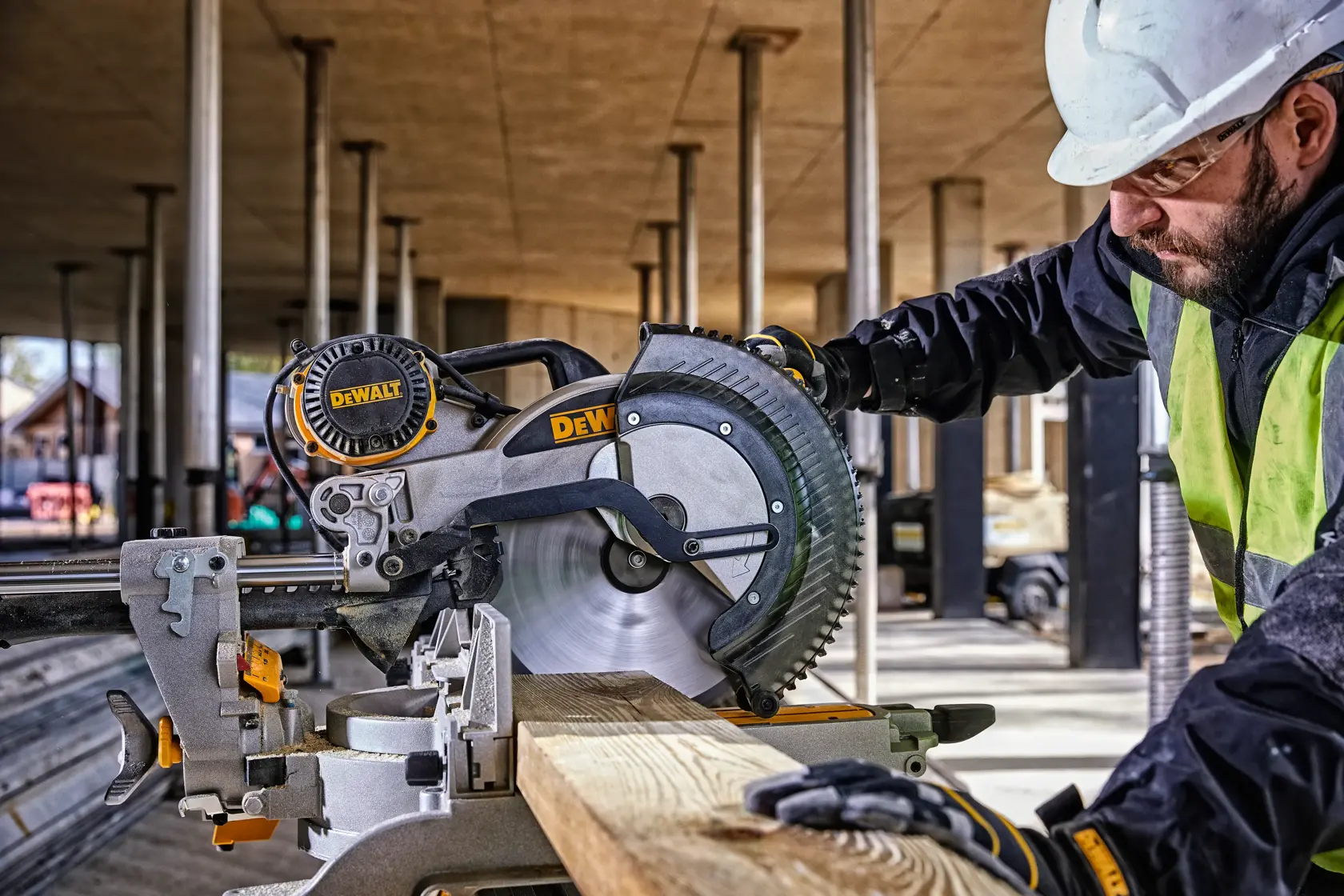 Une personne portant un casque de sécurité blanc et des gants utilise une scie à onglet jaune DEWALT DWS780 sur un chantier de construction. À l’arrière-plan, on voit la structure inachevée d’un bâtiment avec des colonnes et des poutres.
