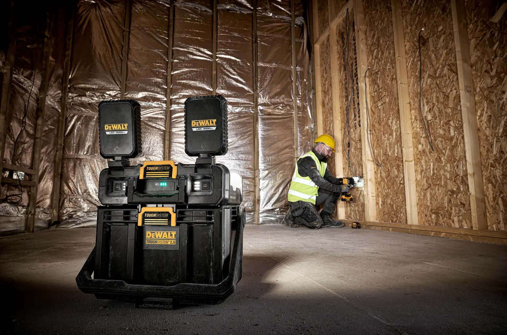 Una radio para obra DEWALT ToughSystem está colocada en el suelo de una construcción con paredes de madera sin terminar y aislamiento. Al fondo, un trabajador lleva casco amarillo y chaleco reflectante.