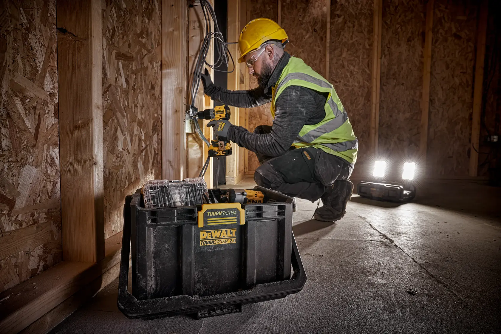Una persona con casco amarillo y chaleco de alta visibilidad utiliza una herramienta eléctrica DEWALT para trabajar en cables dentro de un sitio de construcción de madera. Una caja de herramientas DEWALT TOUGHSYSTEM 2.0 está en primer plano y las luces portátiles iluminan la zona.