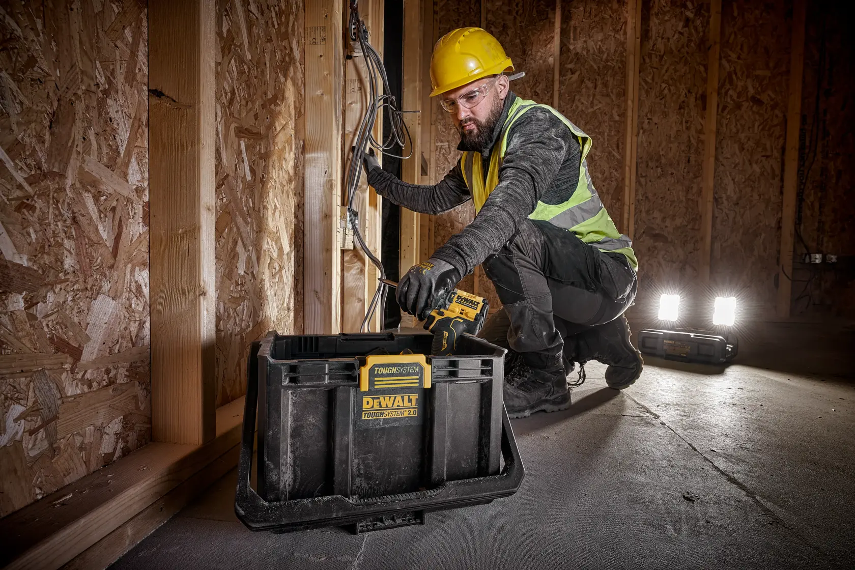 Una persona con casco amarillo y chaleco de seguridad está trabajando con cables eléctricos dentro de un edificio de estructura de madera. Enfrente tiene una caja de almacenamiento DEWALT ToughSystem, y hay dos lámparas portátiles en el suelo.
