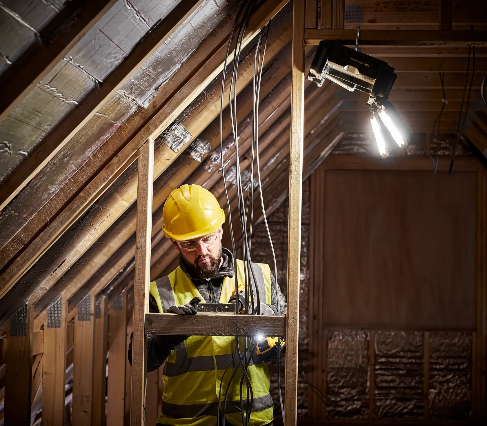 Un trabajador de la construcción con casco amarillo y chaleco reflectante está trabajando en un ático parcialmente terminado. Maneja herramientas y cables eléctricos, y una luz de trabajo portátil DEWALT está instalada arriba, iluminando la zona.