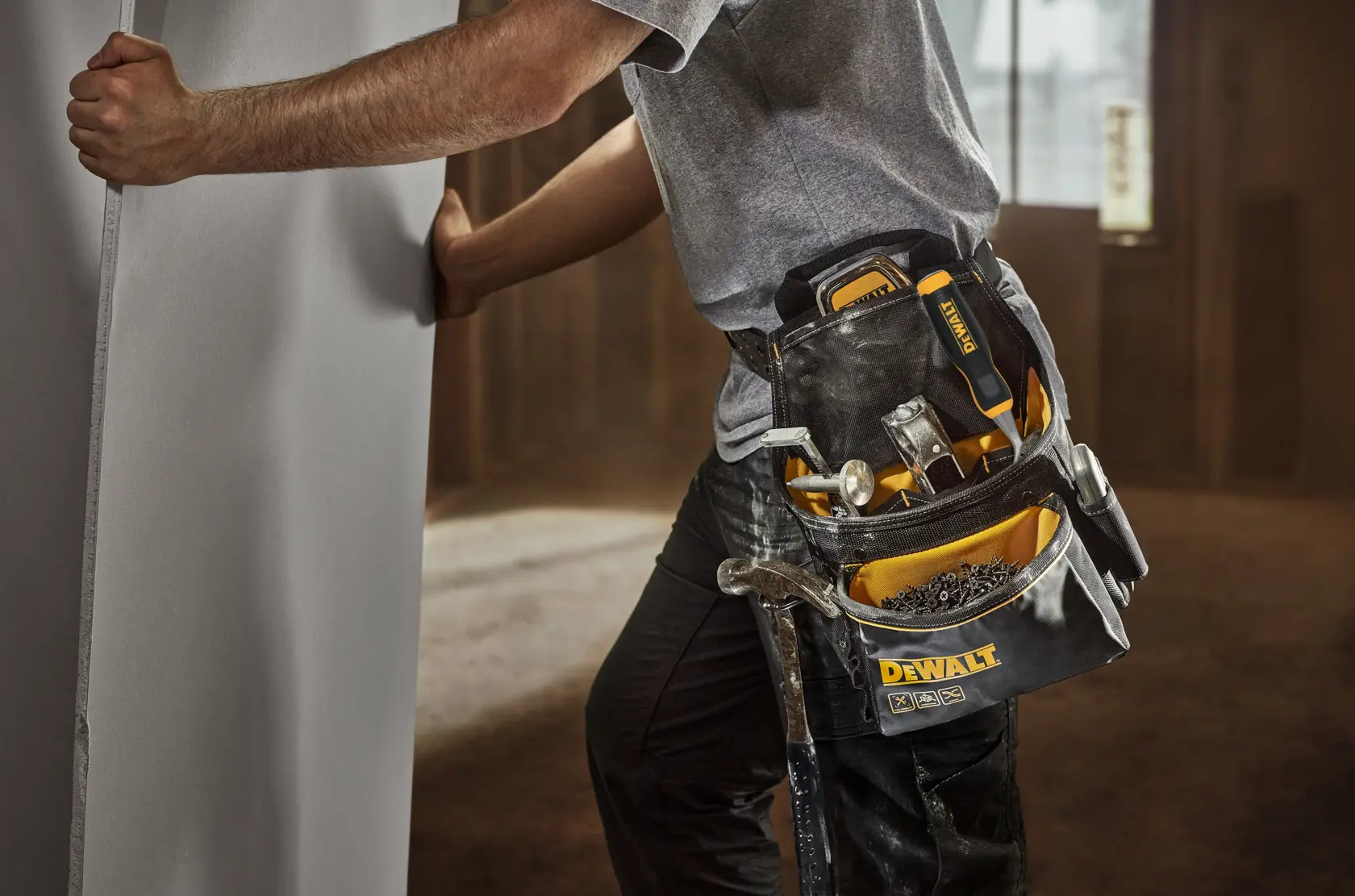 Una persona con un cinturón de herramientas DEWALT sostiene un panel grande. El cinturón lleva varias herramientas manuales, incluido un martillo, destornilladores y fijaciones. La escena está ambientada en interiores, sugiriendo un entorno de construcción o instalación.