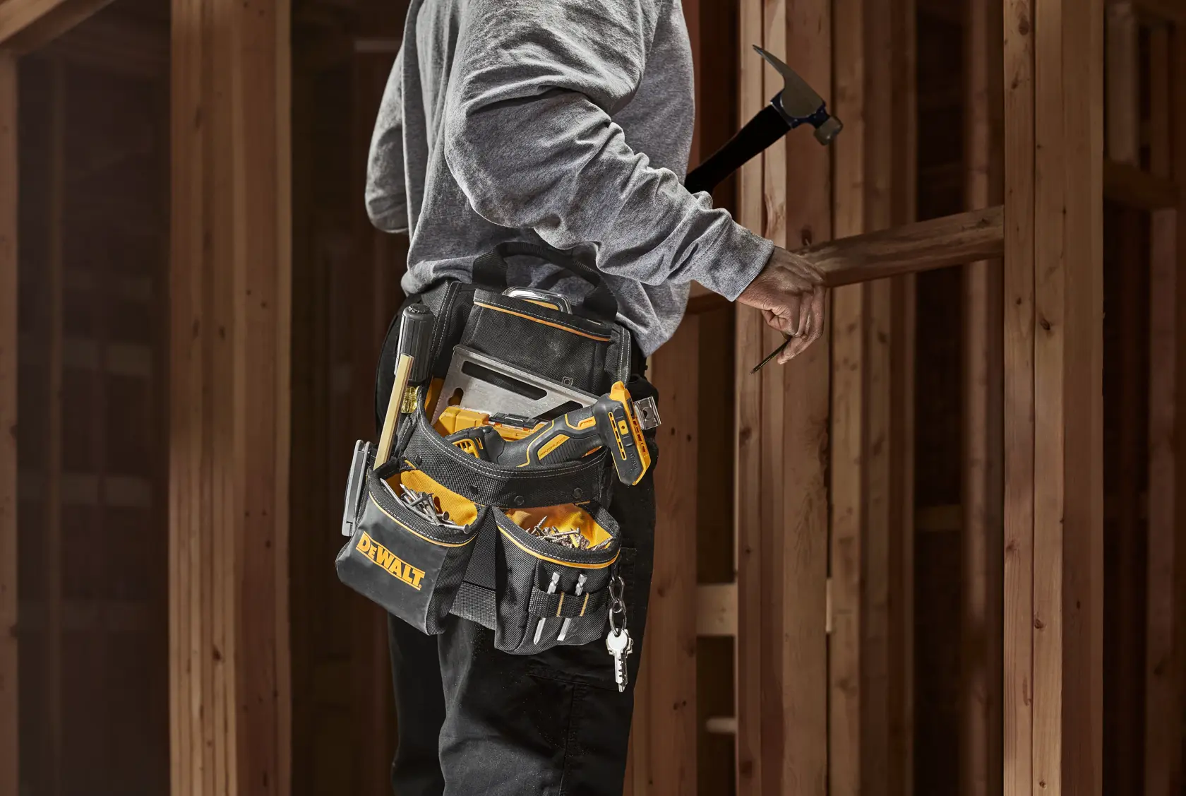 Una persona lleva un cinturón de herramientas DEWALT lleno de varias herramientas manuales y trabaja en una estructura de madera. El cinturón tiene múltiples bolsillos y compartimentos para guardar las herramientas de manera organizada.