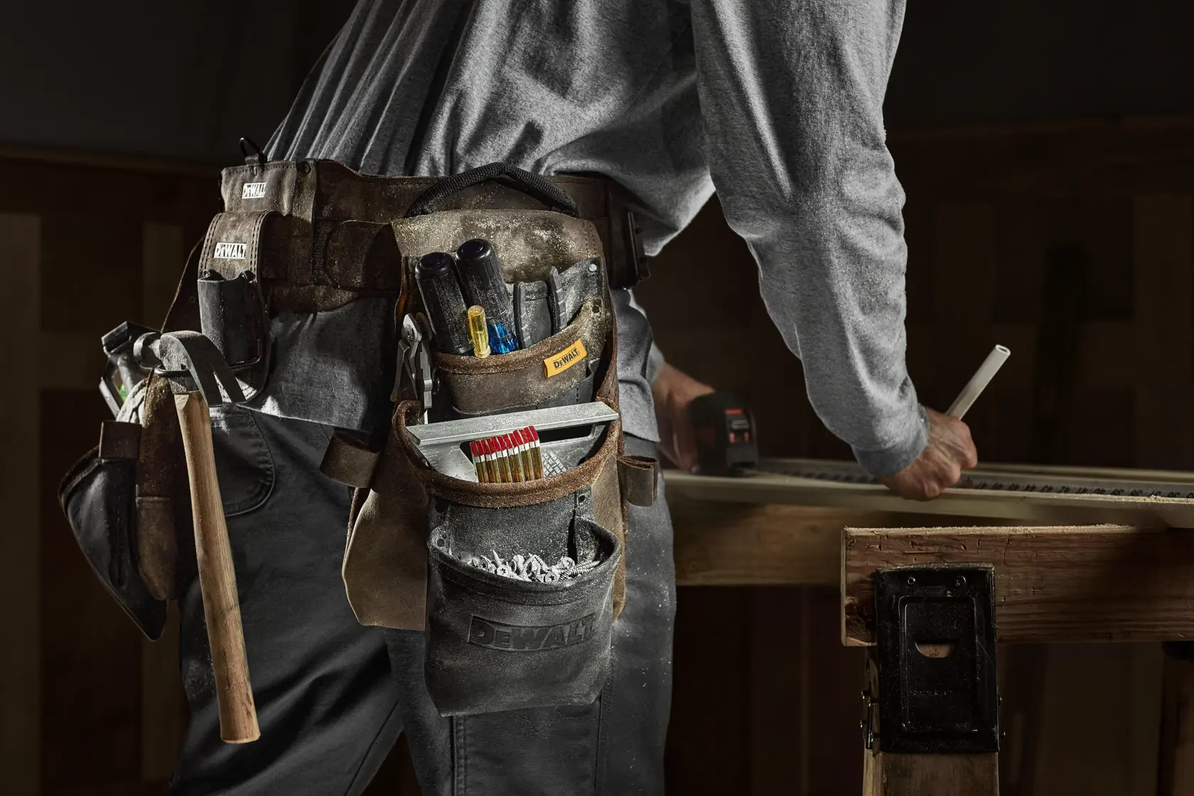 Nahaufnahme einer Person, die einen DEWALT-Werkzeuggürtel mit verschiedenen Werkzeugen wie Hammer, Bleistiften und Schraubendrehern trägt. Die Person arbeitet an einem Holztisch und zeichnet auf einem Holzstück.