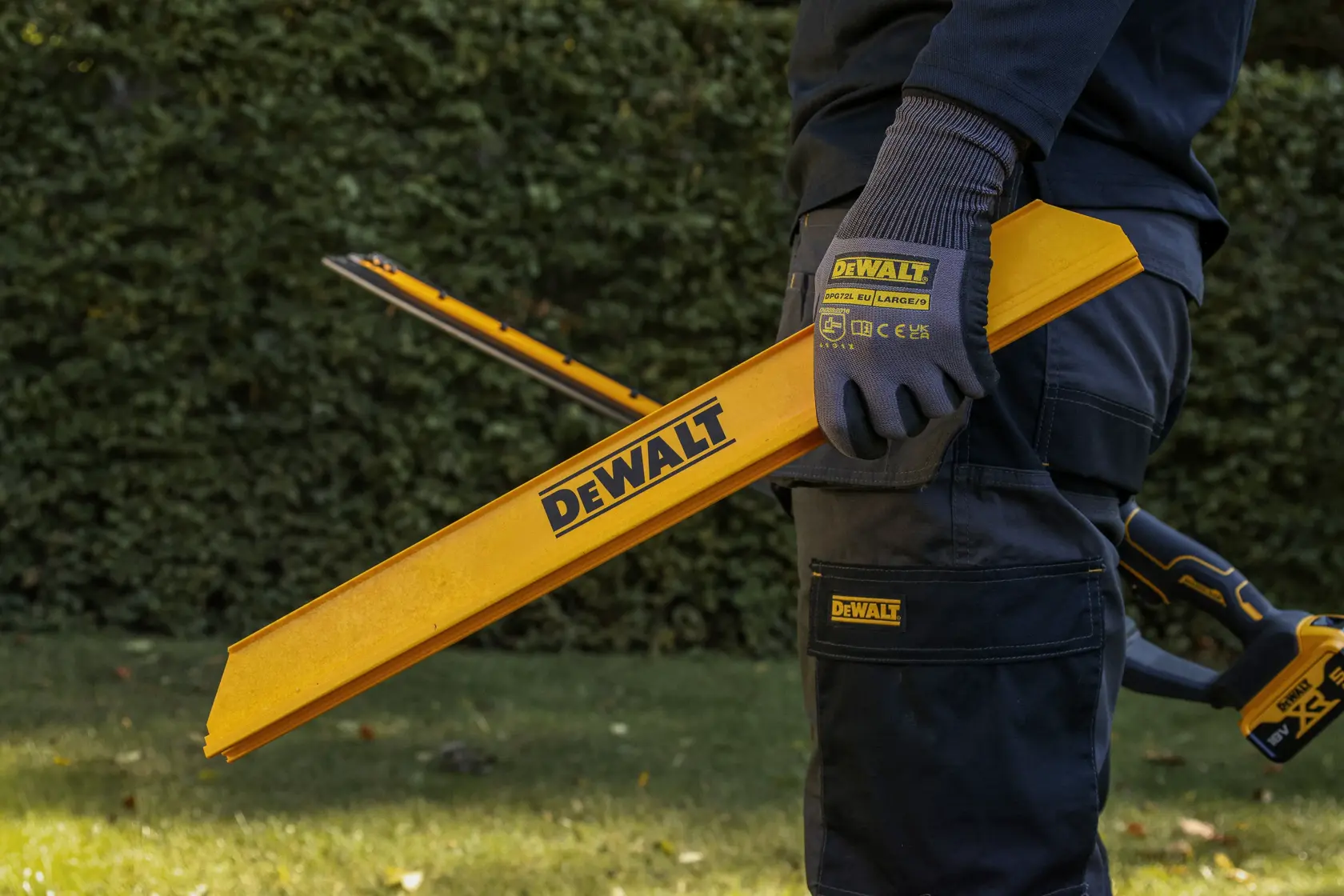 Personne dehors tenant un outil jaune DEWALT, gants et pantalon de travail. Haie verte derrière.
