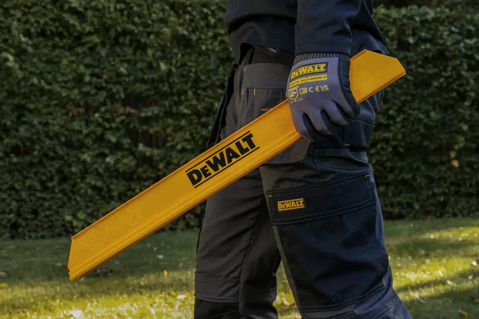 Personne tenant un outil jaune DEWALT dehors, portant des gants et un pantalon DEWALT.